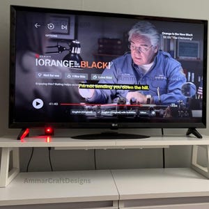 May include: A flat-screen television displaying a scene from "Orange is the New Black" on a white table. The table has a rectangular top and angled legs. A red light is visible on the table.