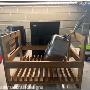 May include: A bamboo dish rack with a stainless steel saucepan. The rack has a rectangular frame with vertical slots for plates and a section for utensils. The saucepan is silver with a long handle. The background includes a black device displaying the time.