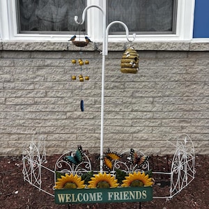 May include: A white metal garden stake with a bird feeder, wind chime, and a decorative beehive. The base features sunflowers, butterflies, and a sign that reads "WELCOME FRIENDS". The AmmarCraftDesigns logo is at the bottom.