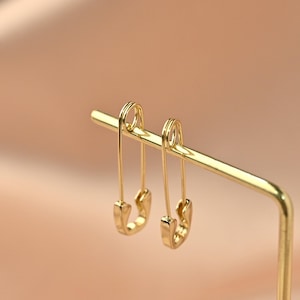 Könnte beinhalten: Ein Paar goldfarbene Sicherheitsnadel-Ohrringe hängen an einem goldenen Präsentationsständer. Die Ohrringe haben die Form von Sicherheitsnadeln, mit einem gebogenen Boden und einem geraden Stift. Der Hintergrund ist eine weiche, neutrale Farbe.