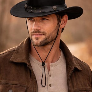 May include: Black cowboy hat with a leather band and decorative studs. The hat has a wide brim and a chin strap. The person in the image is wearing a brown jacket and a light-colored shirt.
