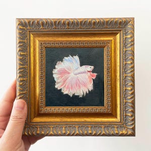 Könnte beinhalten: Gerahmtes Gemälde eines Kampffisches. Der Fisch hat rosa, weiße und blaue Flossen vor einem dunklen Hintergrund. Der Rahmen ist mit einer goldenen Oberfläche verziert. Das Kunstwerk wird von einer Hand gehalten.