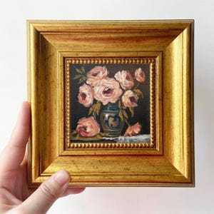 May include: A framed miniature painting of pink roses in a blue and white vase. The painting is set against a dark background and framed in a gold-colored, ornate frame. The artwork is held by a hand.