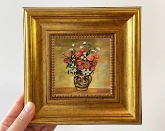 Puede incluir: Una pequeña pintura al óleo cuadrada en un marco dorado grueso y ornamentado. La obra representa un ramo de flores rojas y blancas dispuestas en un jarrón oscuro con patrones sobre un fondo suave amarillo y marrón con pinceladas visibles.