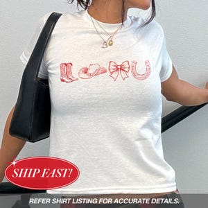 May include: A white short-sleeved t-shirt featuring a red graphic print across the chest. The design consists of a pair of cowboy boots, a cowboy hat, a decorative bow, and a horseshoe, all rendered in a detailed, sketch-like line art style.