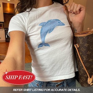 Könnte beinhalten: Ein weißes Kurzarm-T-Shirt mit einem blauen Delfin-Motiv, das mittig auf der Brust aufgedruckt ist. Die Delfin-Illustration hat ein strukturiertes, gesprenkeltes Aussehen in hellen und mittleren Blautönen und ist in einer Sprungbewegung dargestellt.