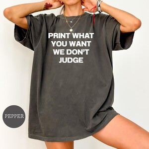 May include: A charcoal grey short-sleeve t-shirt featuring the white text PRINT WHAT YOU WANT WE DON'T JUDGE centred on the chest. The garment has a crew neckline and a relaxed fit. A small circular icon in the corner displays the word PEPPER.