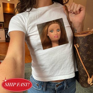 Könnte beinhalten: Weißes T-Shirt mit rundem Ausschnitt und einem quadratischen Bild eines Puppengesichts mit überraschtem Ausdruck. Die Person trägt eine blaue Jeans und eine braune Handtasche. Nahaufnahme.