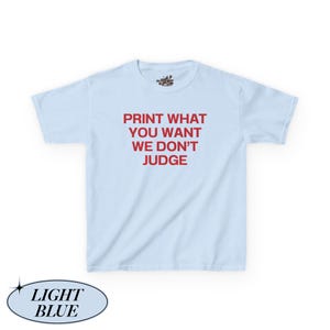 May include: Light blue t-shirt with the red text "PRINT WHAT YOU WANT WE DON'T JUDGE". The shirt has short sleeves and a crew neck. The shirt is a light blue color.