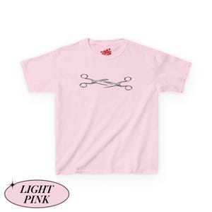May include: Pale pink t-shirt featuring a graphic of two crossed scissors. The t-shirt has a crew neck and short sleeves. The text "LIGHT PINK" is in a light pink oval at the bottom of the image. A small logo is at the top of the shirt.