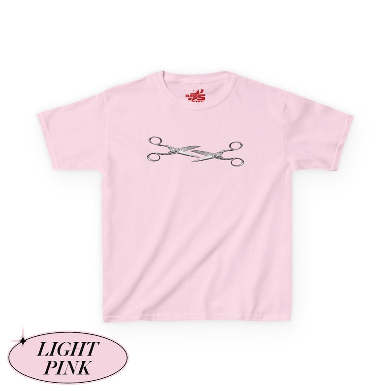May include: Pale pink t-shirt featuring a graphic of two crossed scissors. The t-shirt has a crew neck and short sleeves. The text "LIGHT PINK" is in a light pink oval at the bottom of the image. A small logo is at the top of the shirt.