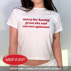 May include: White t-shirt with the text "sorry for having great tits and correct opinions" in red. The shirt has short sleeves and a crew neck. A red and white oval graphic with the words "SHIP FAST!" is at the bottom.