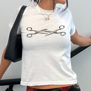 May include: White t-shirt with a black graphic of two scissors. The scissors are facing each other. The shirt has short sleeves and a crew neck. A black shoulder bag is visible.
