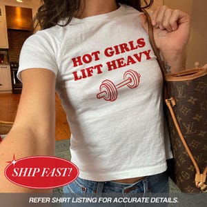 Könnte beinhalten: Ein weißes kurzärmeliges T-Shirt mit dem roten Schriftzug "HOT GIRLS LIFT HEAVY" über einer roten Grafik einer Hantel. Das T-Shirt wird zu einer blauen Jeans getragen und mit einer braunen, gemusterten Umhängetasche kombiniert.