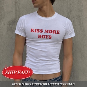 Könnte beinhalten: Ein weißes kurzärmeliges T-Shirt mit dem Text KISS MORE BOYS, der in roten Großbuchstaben auf der Brust gedruckt ist. Das Kleidungsstück hat einen Rundhalsausschnitt und eine körpernahe Passform aus schlichtem weißem Stoff mit roter Schrift.