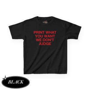 May include: Black t-shirt with the text "PRINT WHAT YOU WANT WE DON'T JUDGE" in red. The word "BLACK" is in a black oval at the bottom left. The shirt is a short-sleeved crew neck.