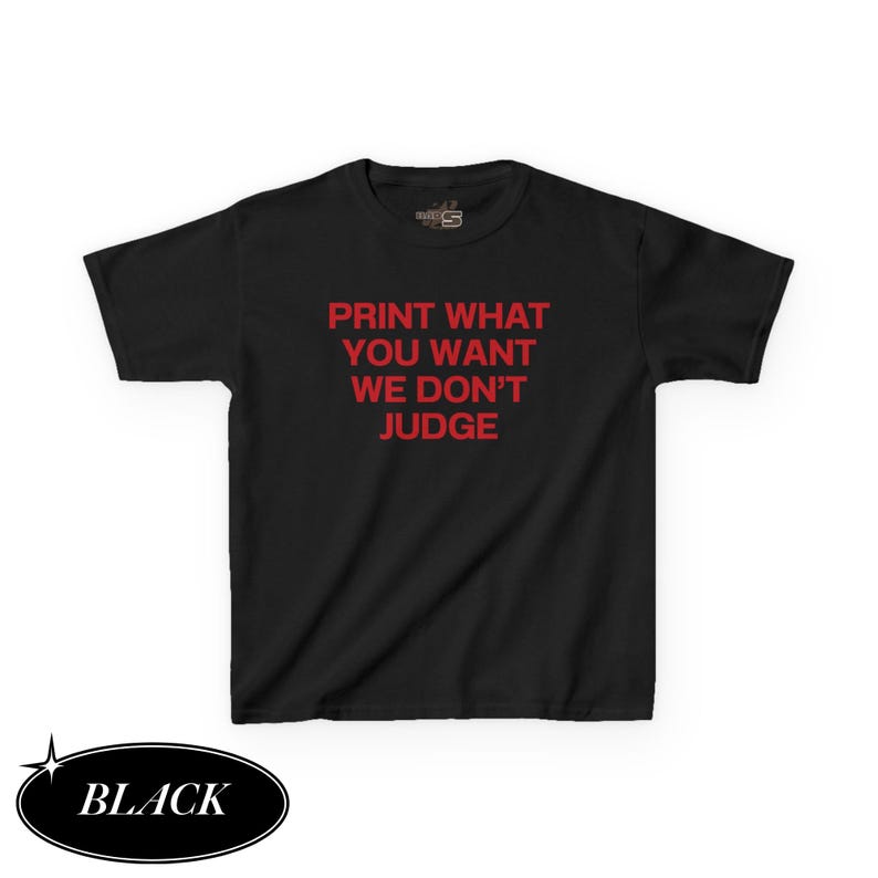 May include: Black t-shirt with the text "PRINT WHAT YOU WANT WE DON'T JUDGE" in red. The word "BLACK" is in a black oval at the bottom left. The shirt is a short-sleeved crew neck.