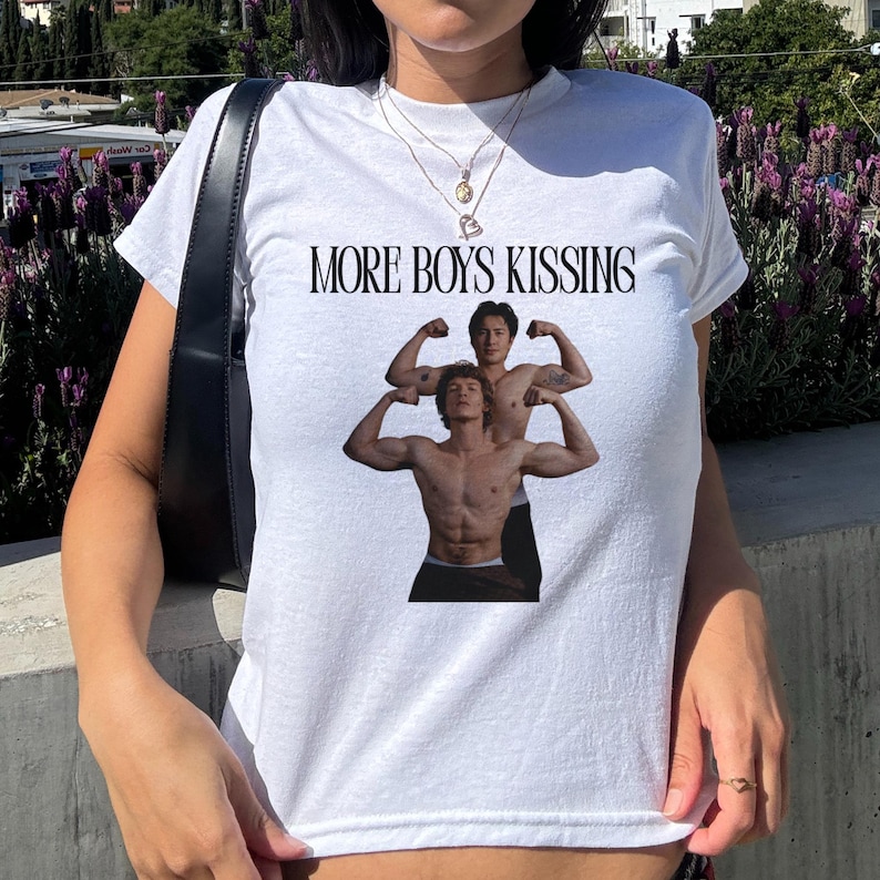 May include: White t-shirt with the text "MORE BOYS KISSING" above a graphic of two shirtless men flexing their biceps. The t-shirt is a crew neck style.
