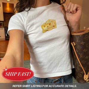 May include: A white short-sleeved t-shirt featuring a central graphic illustration of a yellow wedge of Swiss cheese with holes. The garment is worn with blue denim jeans and a brown patterned shoulder bag with gold-toned hardware details.