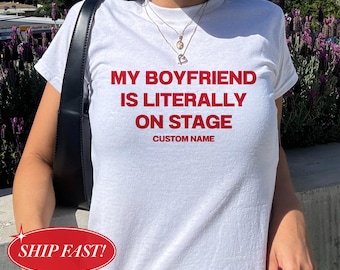 May include: White t-shirt with the red text "MY BOYFRIEND IS LITERALLY ON STAGE" and "CUSTOM NAME" below. The shirt is a crew neck style. A black shoulder bag is visible on the left side. The bottom of the image has the text "REFER SHIRT LISTING FOR ACCURATE DETAILS."