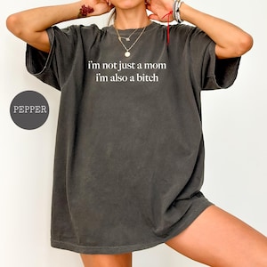 May include: Dark grey t-shirt with the text "I'm not just a mom I'm also a bitch" in white. The shirt is a relaxed fit and has short sleeves. A round label with the word "PEPPER" is on the left side.