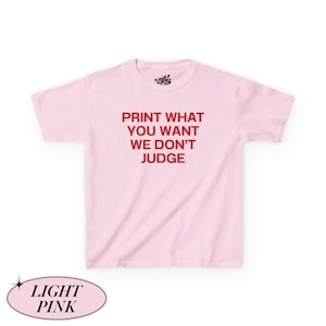 May include: Light pink t-shirt with the text "PRINT WHAT YOU WANT WE DON'T JUDGE" in red. The shirt has short sleeves and a crew neck. The words "LIGHT PINK" are in a light pink oval at the bottom.