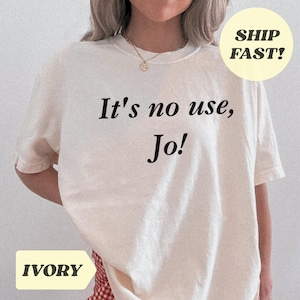 May include: Cream-coloured t-shirt featuring the phrase "It's no use, Jo!" in black script. The shirt has short sleeves and a round neckline. A yellow circle with the words "SHIP FAST!" is in the upper right corner. The word "IVORY" is in a yellow rectangle at the bottom.