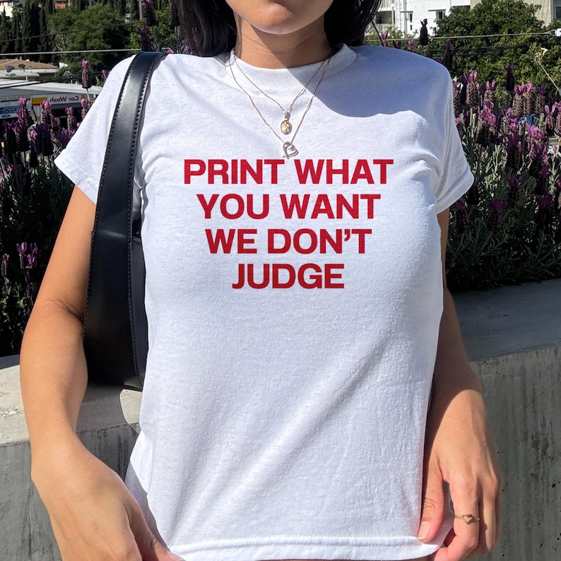 May include: White t-shirt with the red text "PRINT WHAT YOU WANT WE DON'T JUDGE". The shirt is short-sleeved and appears to be made of cotton. A black shoulder bag is visible on the left side of the image.