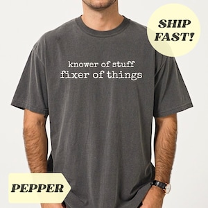 Könnte beinhalten: Dunkelgraues T-Shirt mit dem weißen Text "knower of stuff fixer of things". Das Shirt hat kurze Ärmel und einen Rundhalsausschnitt. Das Wort "PEPPER" steht in einem gelben Banner unten links.
