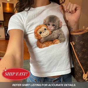 以下が含まれることがあります： 2匹の猿が抱き合っているグラフィックが特徴の白いTシャツ。1匹の猿は茶色、もう1匹はグレーです。画像の下部にあるテキストは「REFER SHIRT LISTING FOR ACCURATE DETAILS」と書かれています。