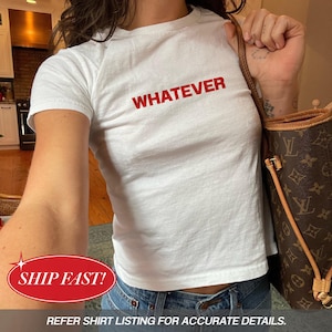 May include: White t-shirt with the word "WHATEVER" printed in red. The shirt is short-sleeved and appears to be made of cotton. The model is wearing blue jeans and carrying a brown handbag.