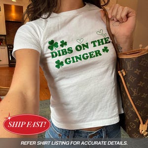 May include: A white t-shirt featuring the green text "DIBS ON THE GINGER", shamrocks, and hearts. The shirt is worn with blue jeans. A brown handbag is also visible in the image.
