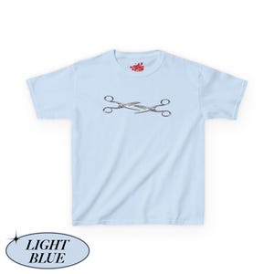 May include: Pale blue t-shirt featuring a mirrored pair of scissors in silver and black. The shirt has a crew neck and short sleeves. The text "LIGHT BLUE" is in a light blue oval at the bottom left.