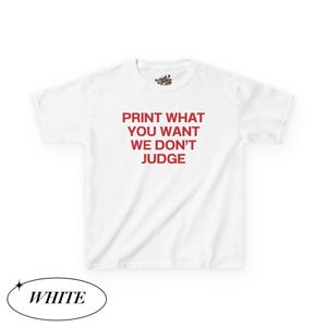 May include: White t-shirt with the red text "PRINT WHAT YOU WANT WE DON'T JUDGE". The shirt has short sleeves and a crew neck. The word "WHITE" is in a black oval at the bottom.