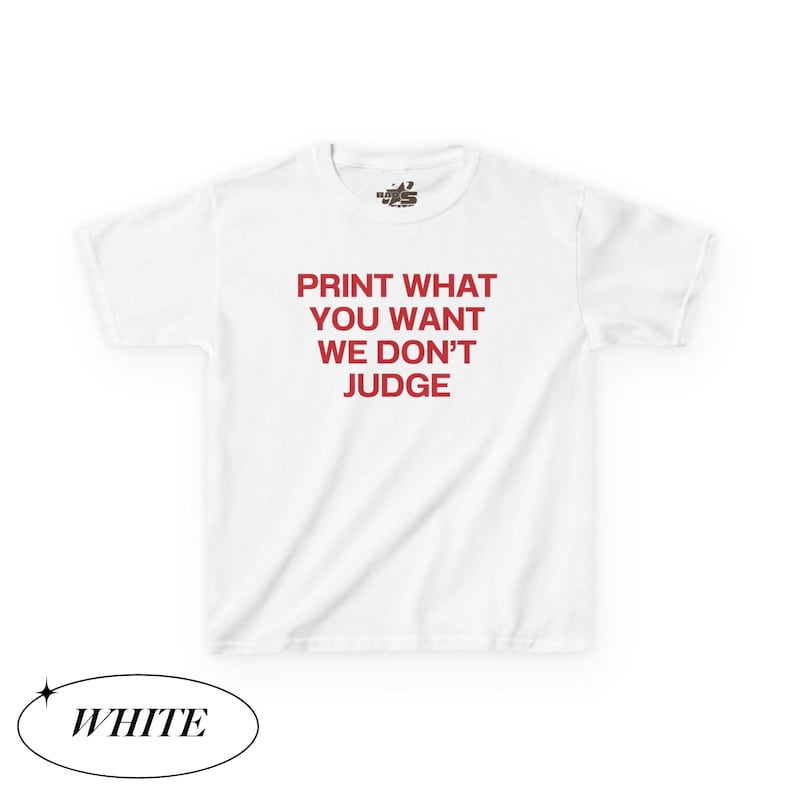 May include: White t-shirt with the red text "PRINT WHAT YOU WANT WE DON'T JUDGE". The shirt has short sleeves and a crew neck. The word "WHITE" is in a black oval at the bottom.