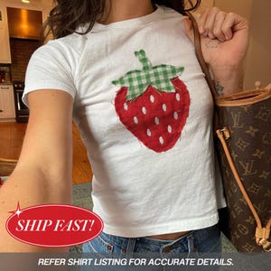 May include: A white short-sleeved t-shirt featuring a large, central strawberry applique. The strawberry is constructed from red fabric with white embroidered seeds and a green and white gingham fabric leaf at the top, showcasing raw edges.