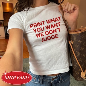 May include: White t-shirt with the red text "PRINT WHAT YOU WANT WE DON'T JUDGE". The shirt is a crew neck style and is paired with blue jeans. A brown handbag is visible in the background.
