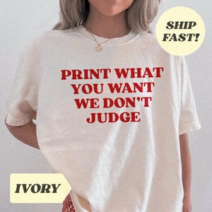Op de afbeelding: Ivoor t-shirt met de rode tekst "PRINT WHAT YOU WANT WE DON'T JUDGE". Het shirt heeft korte mouwen en een ronde hals. Een gele cirkel met "SHIP FAST!" staat rechtsboven.