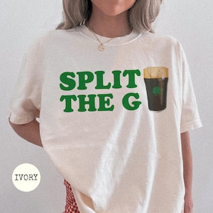 以下が含まれることがあります： アイボリー色のTシャツ。緑色の文字で「SPLIT THE G」と書かれており、シャムロックの付いたスタウトのパイントのグラフィックが描かれています。シャツはクルーネックで半袖です。「IVORY」の文字が下に印刷されています。