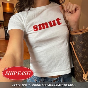 May include: A white short-sleeved t-shirt featuring the word "smut" printed in bold red lowercase letters across the chest. The shirt has a crew neckline and a standard fit, paired with a brown patterned shoulder bag with gold-tone hardware.