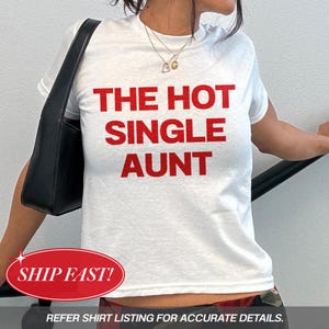 May include: A white short-sleeved t-shirt featuring the text THE HOT SINGLE AUNT printed in bold red capital letters across the chest. The shirt is paired with a black shoulder bag and a gold-toned necklace with a heart-shaped pendant.