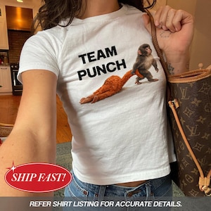 以下が含まれることがあります： 「TEAM PUNCH」の黒い文字が入った白いTシャツ。Tシャツには、ふわふわのオレンジ色の生き物を引っ張る猿のグラフィックが描かれています。Tシャツはブルージーンズと組み合わせています。