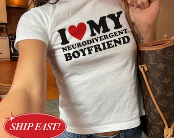 Peut inclure: Un t-shirt blanc à manches courtes avec le texte noir I LOVE MY NEURODIVERGENT BOYFRIEND, où un cœur rouge remplace le mot LOVE. Le t-shirt est porté avec un jean bleu et accompagné d'un sac à bandoulière marron à motifs.