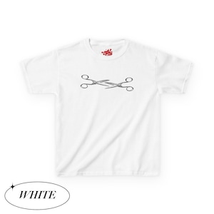 May include: White t-shirt featuring a black and white illustration of crossed scissors. The shirt has a crew neck and short sleeves. The word "WHITE" is in a white oval at the bottom of the image.