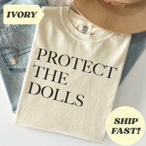 May include: Ivory-colored t-shirt with the text "PROTECT THE DOLLS" in black. The shirt is folded on top of blue jeans, with a tan hat and a silver necklace visible. The shirt has a Comfort Colors tag.