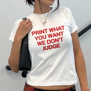 May include: White t-shirt with the red text "PRINT WHAT YOU WANT WE DON'T JUDGE." The shirt has short sleeves and a crew neck. A black purse is visible on the left side of the image.