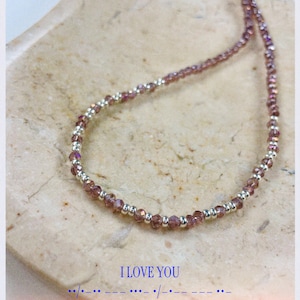 Könnte beinhalten: Eine zarte Halskette mit kleinen, facettierten, lila Perlen und silberfarbenen Perlen. Die Halskette wird auf einer hellen, strukturierten Oberfläche präsentiert. Der Text "I LOVE YOU" und Morsecode sind sichtbar.
