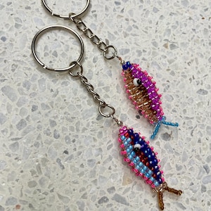 Könnte beinhalten: Zwei Schlüsselanhänger mit Fischperlen. Ein Fisch ist hauptsächlich rosa und goldfarben, der andere blau und rot. Beide haben silberne Schlüsselringe und Ketten. Die Fische haben kleine Perlendetails für Augen und Flossen.