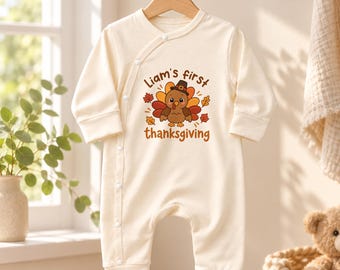 May include: A cream-colored long-sleeve baby romper featuring a graphic of a turkey wearing a pilgrim hat surrounded by autumn leaves. The text reads Liam's first thanksgiving. The garment has a side-snap closure with white buttons.