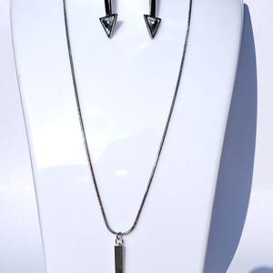 May include: A silver-coloured jewellery set comprising a necklace and matching earrings. The necklace has a fine chain with an arrow-shaped pendant. The earrings are also arrow-shaped. Each arrow has a clear, triangular crystal at the tip.